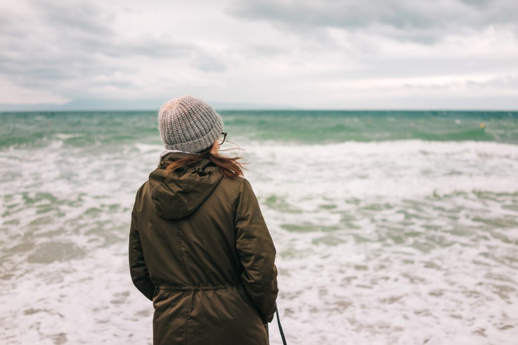 A person standing on a beach, looking out at the ocean with waves crashing. They are wearing a cozy knit hat and a green jacket.