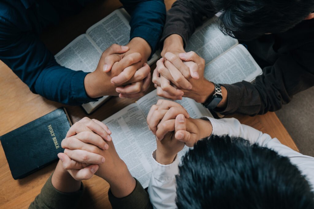 Group of people praying together, holding a Bible and displaying prayer hands in a moment of unity and devotion.