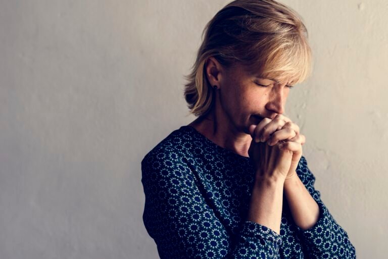 A woman with short blonde hair, eyes closed, and hands clasped together, appearing deep in thought or prayer.